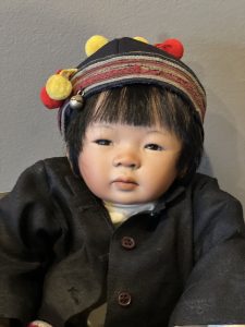 A porcelain doll with black hair and a striped hat, representing a child from China, with a visible red and yellow tie on the hat.