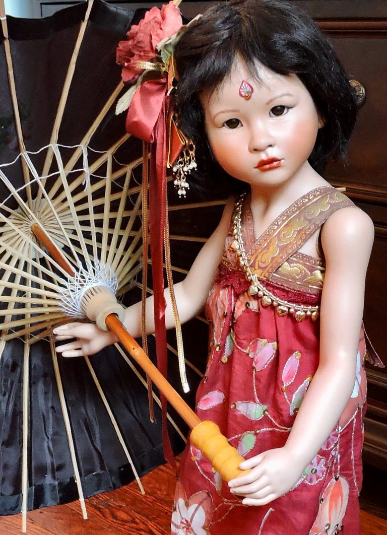 A porcelain doll with a red silk dress and leather sandals, holding a black bamboo umbrella, with a decorative floral hairpin and glass eyes.