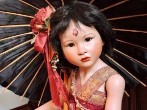 A porcelain doll with a red silk dress and leather sandals, holding a black bamboo umbrella, with a decorative floral hairpin and glass eyes.