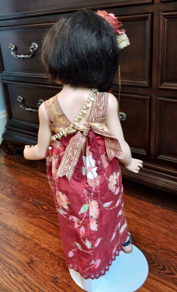A porcelain doll with a red silk dress and leather sandals, holding a black bamboo umbrella, with a decorative floral hairpin and glass eyes.