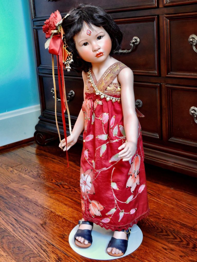 A porcelain doll with a red silk dress and leather sandals, holding a black bamboo umbrella, with a decorative floral hairpin and glass eyes.