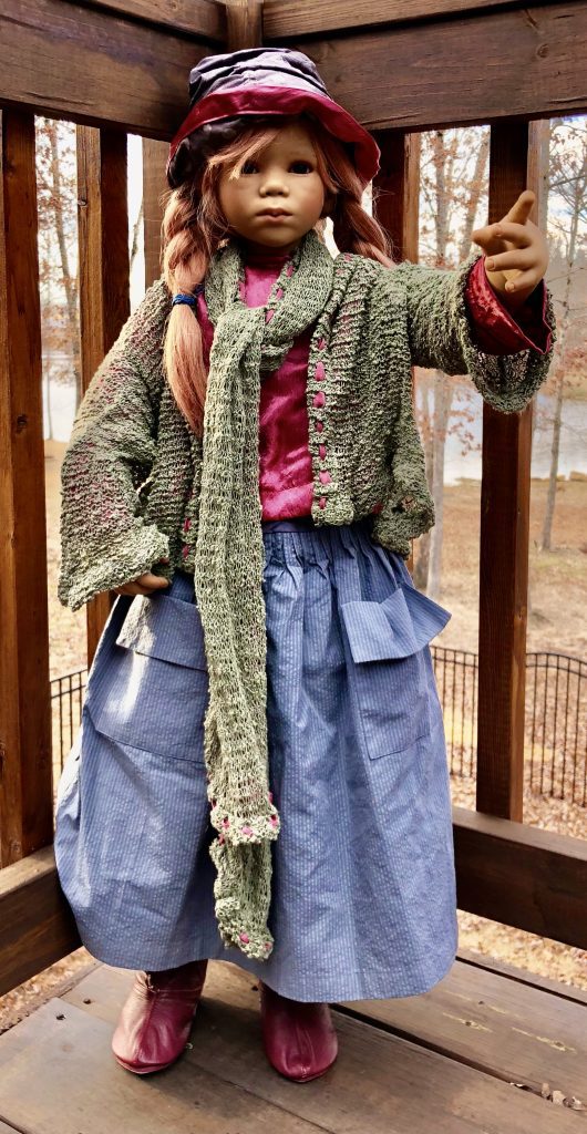 A doll with red hair and wearing a green and red outfit, including a hat and a scarf. The doll has a wooden head and is positioned against a fence.