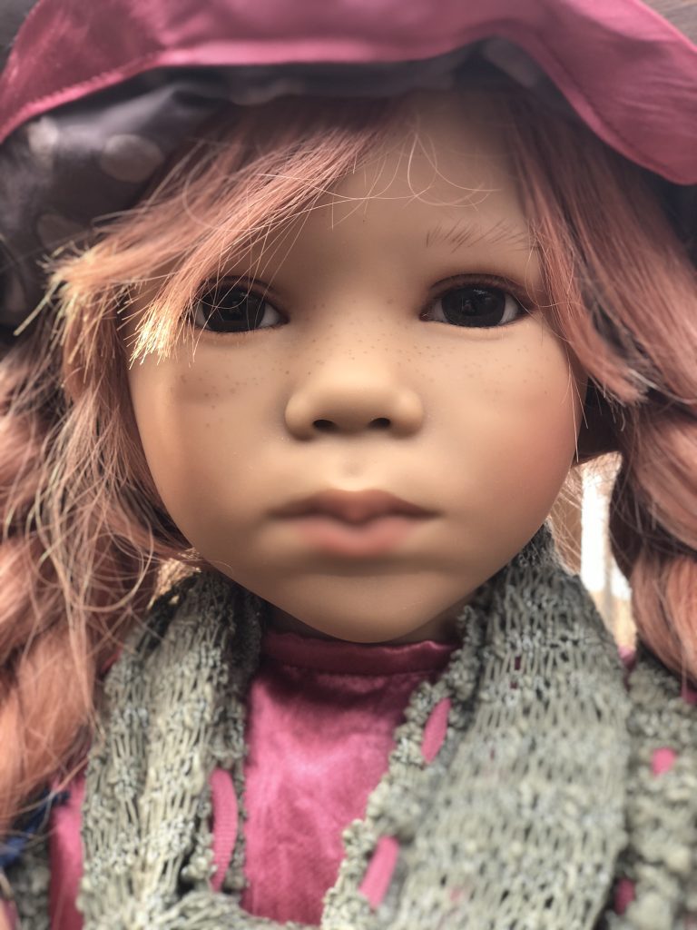 A doll with red hair and wearing a green and red outfit, including a hat and a scarf. The doll has a wooden head and is positioned against a fence.