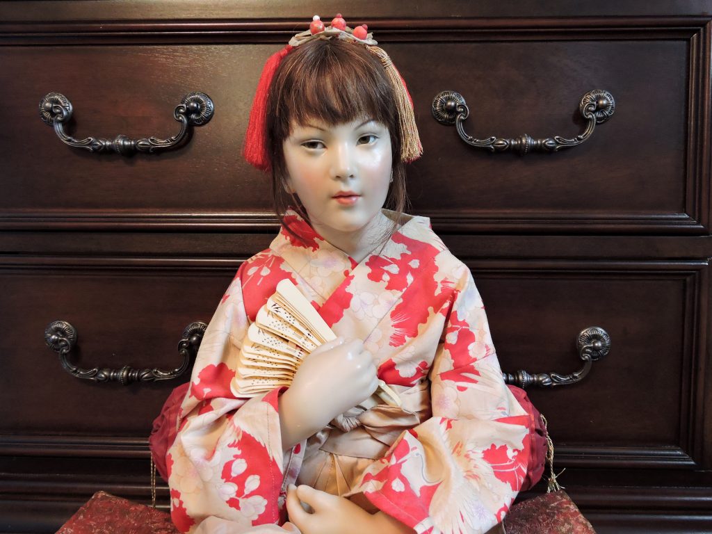 A handcrafted Japanese girl doll made of high-fired clay with a human hair hand-knotted wig, wearing a traditional Japanese kimono with red and gold floral patterns, seated on a mattress-like surface.