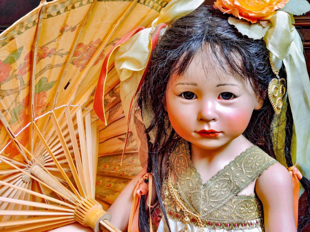 A highly detailed image of a Sakia porcelain doll, featuring a human hair wig, glass eyes, and a silk outfit with a red and gold belt. The doll has a realistic appearance with a natural bamboo umbrella.